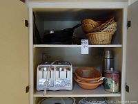 Cabinet shelf showing silver four-slice toaster, ceramic bowls, stainless steel thermos, decorative floral canister, and stacked wicker baskets.