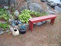 Wide view showing the garden bench alongside a group of various resin frogs, an old metal kettle, some pots, and part of the landscaped garden with plants and rocks.