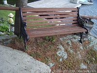 Full front and side view of the wooden slatted bench with decorative cast iron ends and legs on outdoor ground area with some moss and weathering.