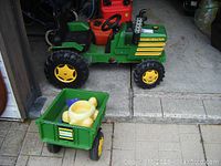 Full view of green and yellow Perego pedal tractor with attached green cart containing a yellow and beige toy.