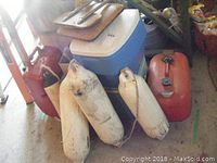 Photo showing three white boat bumpers with rope, two red gas tanks, blue and white cooler, brown boat seat, and vintage metal cooler in background