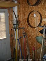 Various garden tools such as rakes, shovels, spades, and tree saws leaning against a wooden wall near a window and door.