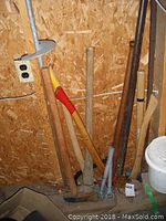 Axes, log roller, pick axe, and other tools with wooden handles leaning against a wooden wall