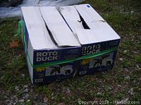 Two original Roto Duck brand boxes containing mechanical duck decoys, secured with green strap, shown outdoors on gravel and grass.