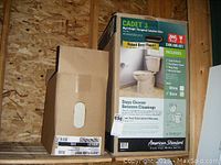 Two boxes showing packaging of American Standard Cadet 3 right height elongated toilet in bone color with details of included parts and specifications.