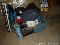 Overview of lot including birdhouse, boxed rotisserie, lawn chairs, cooler, and other items stacked on floor.