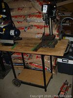 Drill press mounted on wooden work bench with metal frame and wheels, surrounded by bags in storage.