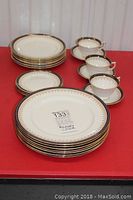 Photo showing stacked Aynsley bone china plates including dinner, salad, and side plates with cups and saucers arranged on side. Pieces have white base with gold floral and cobalt blue border.
