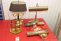Photo showing three lamps on a red table, including hexagonal glass and brass table lamp, brass piano lamp, and smaller table lamp with dark shade.