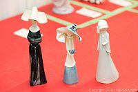 Three porcelain figurines on red surface, front and side views visible, showing styles and colors, with background showing more figurines blurred.