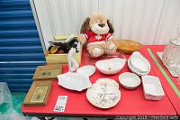 Lot displayed on red table including framed sports medals, white ceramic dishes and bowls, floral scalloped plate, plush dog toy, wooden basket, glass relish dish, box of clothes brushes, and magnifying glass.