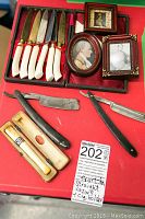 Photo showing two antique straight razors, six fruit knives with white handles in a box, vintage cigarette holder, three framed portraits on a red background.