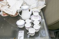 Full espresso coffee set shown on glass table including coffee pot, sugar bowl, creamer, six cups and saucers with white tissue paper in background.