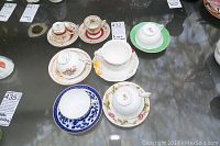 Seven bone china cups and saucers arranged on a table with visible labels indicating lot and item numbers.