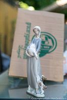 Front view of Lladro figurine showing woman holding a goose and small dog at her feet with box in background.