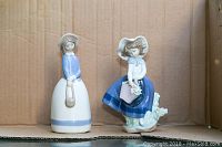 Photo of two porcelain figurines, one in blue and white dress with a bonnet and purse, the other sitting with a basket of flowers in blue skirt and white blouse.