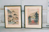 Pair of framed Asian paintings side by side leaning against a door, showing both artworks with matted beige surrounds inside black frames.