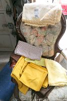 Various linens including cream-colored tablecloth with detailed lace edges, yellow placemats, batik napkins, and a vintage dress on a floral upholstered chair.