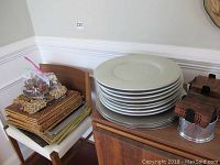 Overall view showing a chair with stacked woven placemats in brown and yellow tones, a bag with fabric napkins or similar, a stack of white ceramic plates, and part of a wooden table.