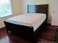 Full view of rustic wood bedframe assembled with mattress. Bedframe shows dark wood with visible grain and rustic finish, includes headboard and footboard.