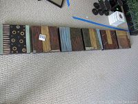 Six square ceramic wall decor pieces laid out in a row on carpet showing varied abstract designs ranging from circular to linear patterns in coordinated muted earth tones.