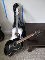 Full view of two parlour-sized acoustic guitars side by side, one black Greg Bennett and one white Crescent, with a black soft case behind against a wall.
