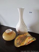 Three decorative items arranged on a dark surface: round amber crackled glass vase, tall white plastic vase with thin brown lines, and a multicolored yellow-rimmed glass piece with abstract undulating shape.