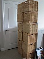 Tall stack of ten wicker baskets in light natural wicker tone with cutout handles, stacked in two columns against a white wall and door.