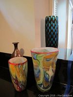 Three vases displayed together: two colorful swirled art glass vases with red rims and one taller metal vase with patina finish, arranged side by side on black surface.