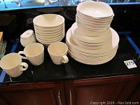 White Crate and Barrel ceramic dishware stacked on black countertop including plates, bowls, mugs, and sugar bowl