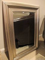 Framed wall mirror standing on the floor, showing the wide silver metallic frame with grooved detailing and beveled mirror glass.