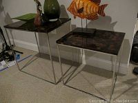 Two metal base end tables with tiled tops, one larger than the other, shown side by side with decorative items on top.