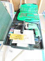 Photo shows the Hitachi corded drill in green with black auxiliary handle inside molded green carrying case along with a large green plastic case containing assorted drill and screwdriver bits, instruction manuals, and a pack of 13 batteries.