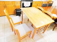 Four wood dining chairs with ladder back design and beige fabric seats around a wooden table in a kitchen with tiled floor. One chair seat fabric is visibly soiled.