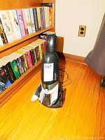 Full view of the Shark cordless vacuum standing on wooden floor by bookshelf, showing upright design and charging base.