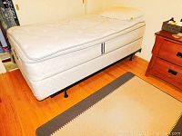 Full side view of the assembled single bed with mattress and box spring, pillow on top, wooden floor and bedside table visible.