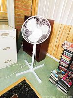 White HomeStyles floor fan standing on a carpeted floor with a cross-shaped base and a power cord, positioned near a filing cabinet and some stacked DVDs.