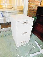 White three-drawer metal filing cabinet shown closed from the front, with visible handle and key lock on top drawer.