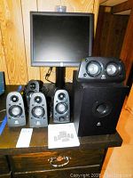 View of the Dell monitor behind the six pieces of Logitech Z506 speaker system including three satellite speakers, one center speaker, one surround speaker and one subwoofer on a black table with wooden paneled background.