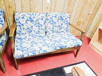 Full view of the loveseat showing the wooden frame and floral cushions against a wood-paneled wall and red floor.