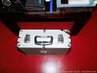 Closed aluminum carry case placed on red floor; black handle and metal latches visible on top