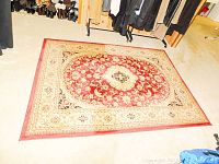 Full view of rectangular carpet with central red medallion and beige border on light carpet floor.