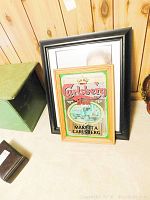 Photo showing Carlsberg Beer mirror in wooden frame leaning against two other framed prints, next to a green box and dark brown small case.