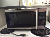 Front view of the Proctor Silex microwave showing the stainless steel front door with black trim and digital keypad control panel on right side, power cord visible.