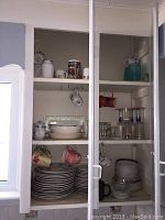 Cabinet interior with stacked ceramic plates, mugs hanging on hooks, clear glasses with purple rims, and serving bowls.