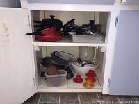 Open kitchen cupboard showing red frying pans, metal baking trays, glass pots with lids, mixing bowls, egg cups, and trivets.