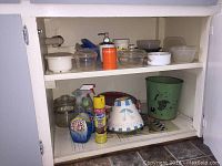 Overview of upper and middle cupboard shelves showing plastic containers, glass dishes, cleaning supplies, orange soap dispenser, paper napkins, and vintage dish with lid alongside a green waste basket below.