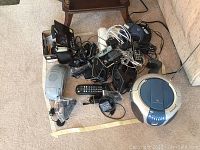 Photo of lot on carpet including a silver and blue CD player, multiple telephones, remotes, power cords, and adapters tangled together.