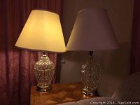 Pair of glass base table lamps on a wooden table with cream-colored shades, one lamp turned on to show working condition.