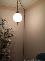 Full side view of hanging lamp illuminated, showing total hanging length of metal chain and cord, and pearl white globe in a room corner near artificial flowers.